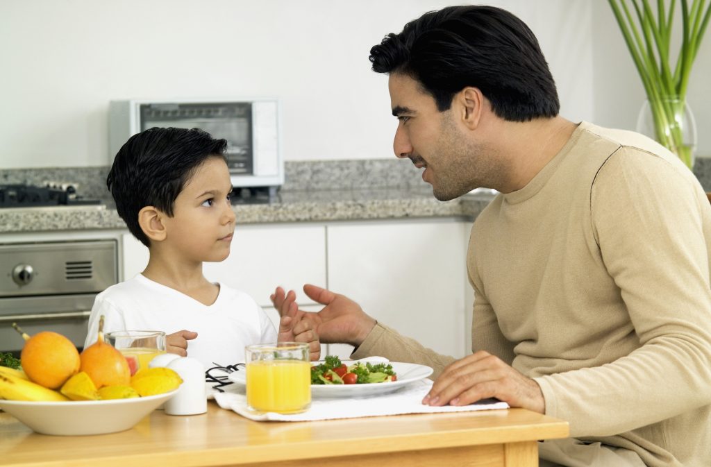 Man and boy sitting at table in kitchen, with salad and orange juice on table and man talking to boy, close-up